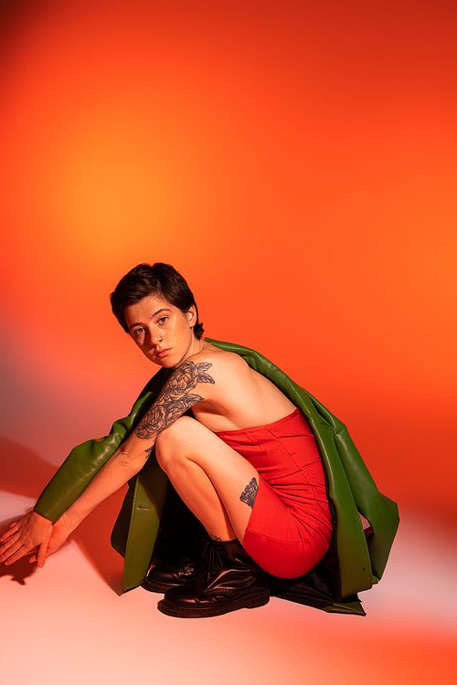 full length of tattooed woman in red corset dress and black boots sitting on haunches and looking at camera on orange and pink background