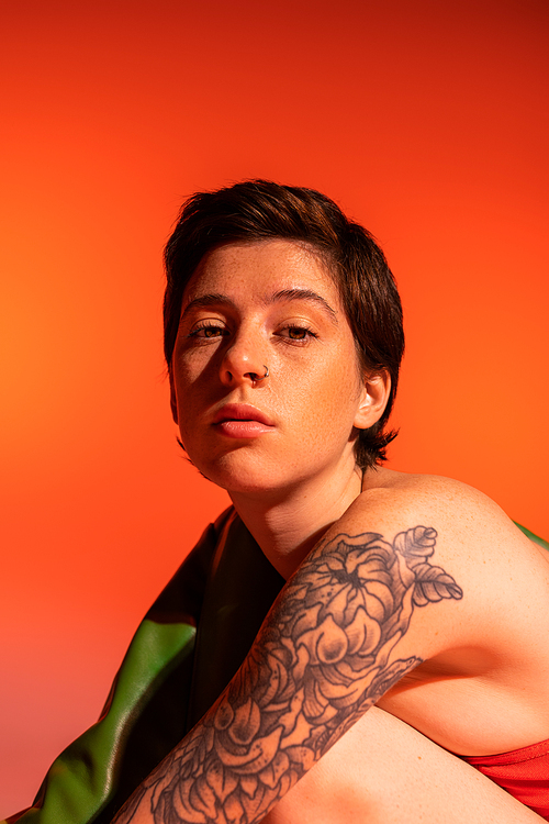 portrait of pensive woman with tattoo and short brunette hair looking at camera on orange background