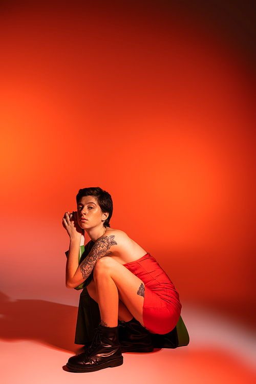 trendy tattooed woman in red strapless dress and black leather boots posing on haunches on orange background