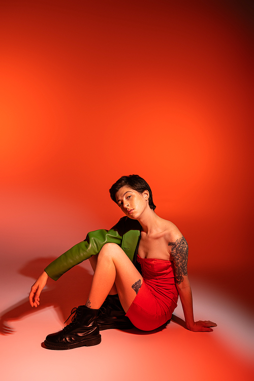 trendy tattooed woman in red strapless dress and black boots looking at camera while posing on orange background