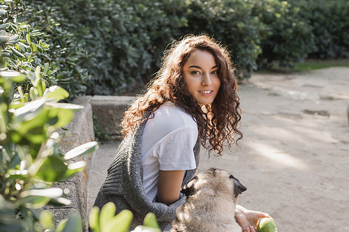 Carefree young curly woman in t-shirt and sweater holding green apple and looking at camera while sitting on stone bench near pug dog and relaxing in park in Barcelona, Spain