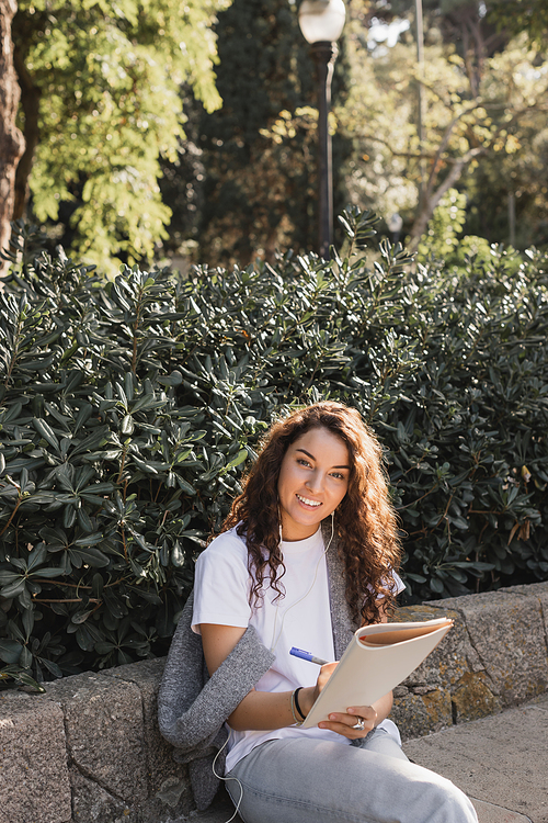 Smiling young and curly woman in casual clothes using wired earphones, writing on notebook and looking at camera while sitting on stone bench in park in Barcelona, Spain