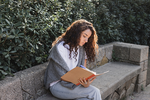 Young and curly brunette woman in t-shirt and sweater writing on notebook while spending time on stone bench near green bushes in park at daytime in Barcelona, Spain