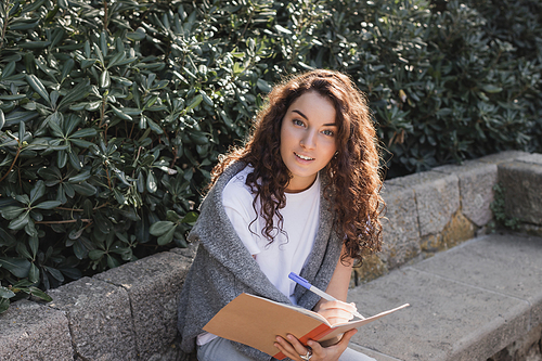 Carefree young and curly woman in sweater and t-shirt looking at camera while writing on notebook and spending time on bench near green bushes in park in Barcelona, Spain