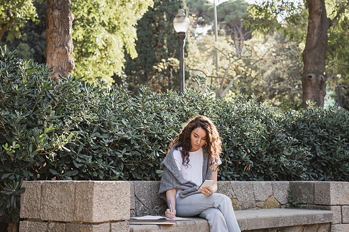 Young and curly freelancer in casual clothes using smartphone and writing on notebook while sitting near laptop on stone bench and green bushes in park in Barcelona, Spain