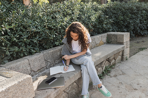 Young and curly woman in warm sweater on shoulders writing on notebook near laptop and smartphone with blank screen on stone bench beside green bushes in park in Barcelona, Spain