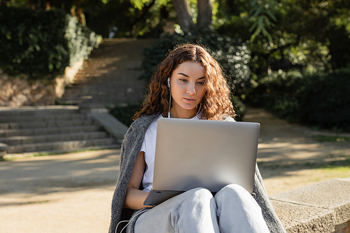 Young and curly freelancer in wired earphones using laptop while sitting on parapet and spending time in blurred green park at daytime in Barcelona, Spain