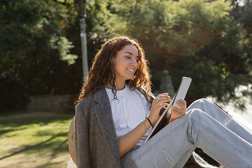 Carefree young and curly freelancer in wired earphones using smartphone and holding laptop while spending time in blurred green park at daytime in Barcelona, Spain