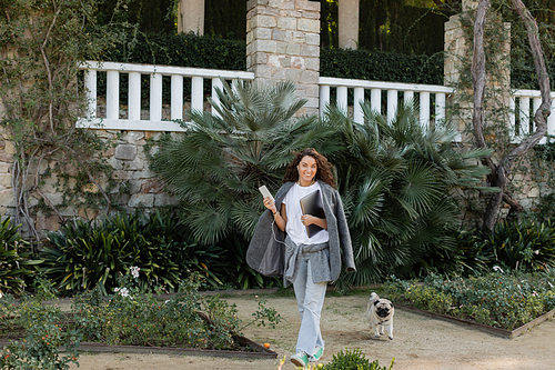 Cheerful and curly freelancer in warm jacket and earphones looking at camera and holding devices while walking near pug dog in park at daytime in Barcelona, Spain