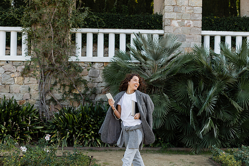 Carefree brunette freelancer in warm jacket listening music in earphones and holding laptop with smartphone while walking in park at daytime in Barcelona, Spain