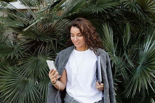 Cheerful young and curly freelancer in warm jacket using earphones and smartphone while holding laptop and standing near green palm trees in park in Barcelona, Spain