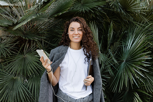 Young and curly freelancer wearing warm jacket and using earphones while holding smartphone and notebook, smiling at camera and standing near palm trees in park in Barcelona, Spain