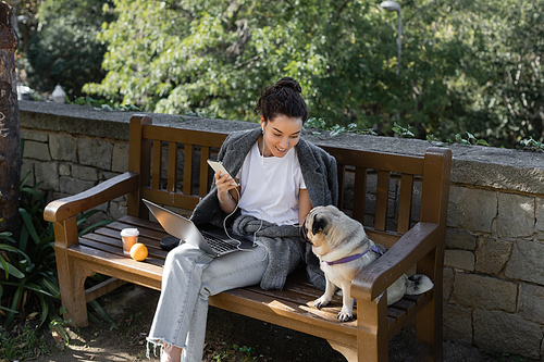 Smiling freelancer in earphones holding smartphone and looking at pug dog while sitting near laptop, coffee to go and orange on wooden bench and spending time in park in Barcelona, Spain