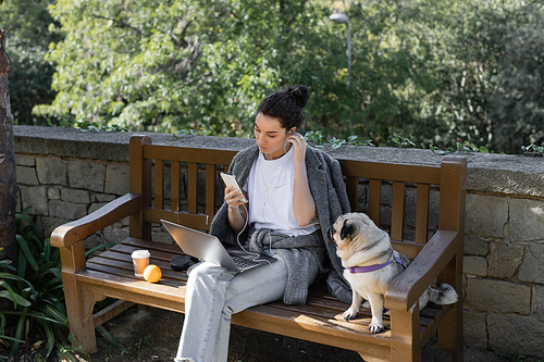 Young and curly freelancer listening music in earphones, using smartphone near laptop and sitting next to pug dog, orange and coffee to go on wooden bench in park in Barcelona, Spain