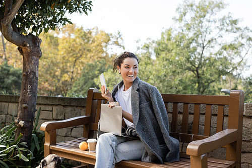 Smiling brunette freelancer in warm jacket using earphones and devices while spending time near pug dog, coffee to go and orange on wooden bench in park in Barcelona, Spain