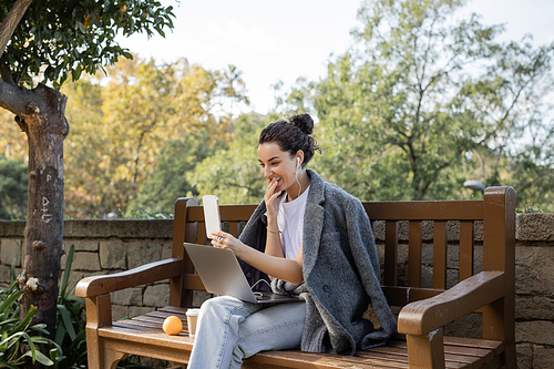 Smiling young freelancer in warm jacket and earphones smiling, covering mouth, having video call on smartphone near laptop and coffee to go on wooden bench in park in Barcelona, Spain