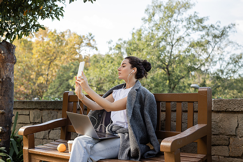 Side view of young freelancer in warm jacket using earphones while having video chat on smartphone near laptop, coffee to go and orange on wooden bench in park in Barcelona, Spain