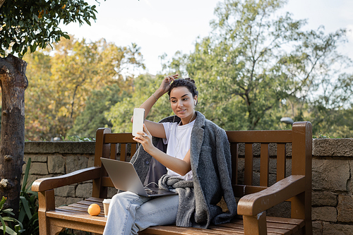 Smiling and young freelancer in warm jacket using earphones and smartphone while sitting near laptop, takeaway drink and fresh orange on wooden bench in park in Barcelona, Spain