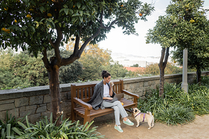 Young brunette freelancer in warm jacket holding coffee to go and using laptop near fresh orange on wooden bench and pug dog near green plants in park in Barcelona, Spain, work from anywhere