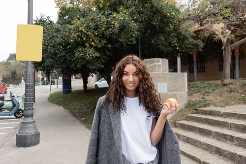 Smiling young and curly woman in warm jacket holding fresh orange and looking at camera while standing on blurred urban street at background in Barcelona, Spain, street lamp, motor scooter