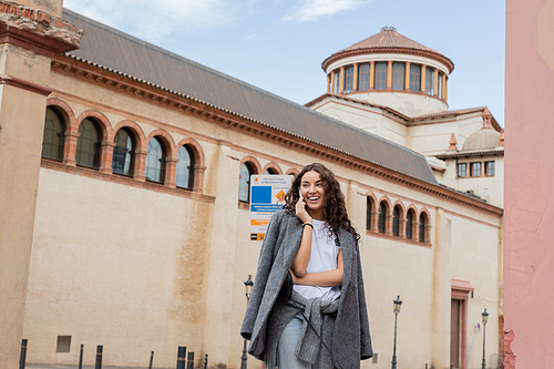 Smiling young and curly woman in warm jacket and casual clothes talking on smartphone and looking away while standing near historic landmark on urban street in Barcelona, Spain, ancient building