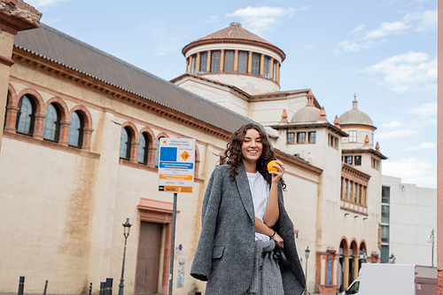 Positive young woman in casual grey jacket holding fresh orange and leash while looking at camera with historical landmark at background on city street in Barcelona, Spain, ancient building