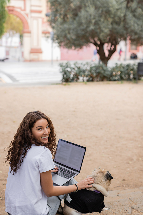 Carefree young and curly freelancer in casual clothes looking at camera while using laptop and petting pug dog on stairs in blurred park in Barcelona, Spain, white t-shirt