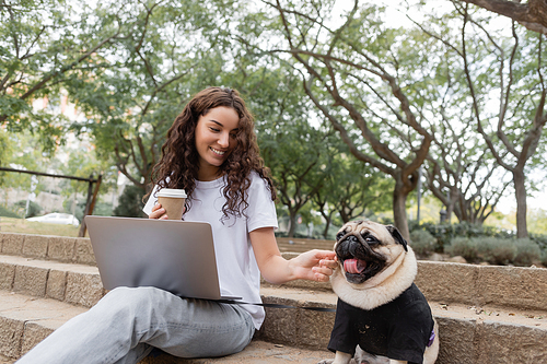 Smiling and curly freelancer in casual clothes holding coffee to go near laptop and petting pug dog while sitting on stairs in blurred park in Barcelona, Spain