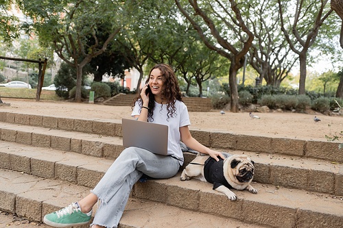 Cheerful brunette freelancer in casual clothes talking on smartphone near laptop and petting pug dog on stairs while spending time in blurred park in Barcelona, Spain