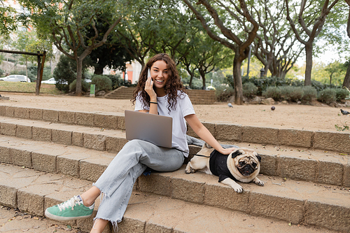 Positive and curly young freelancer talking on smartphone near laptop, looking at camera and petting pug dog while sitting on stairs in park in Barcelona, Spain