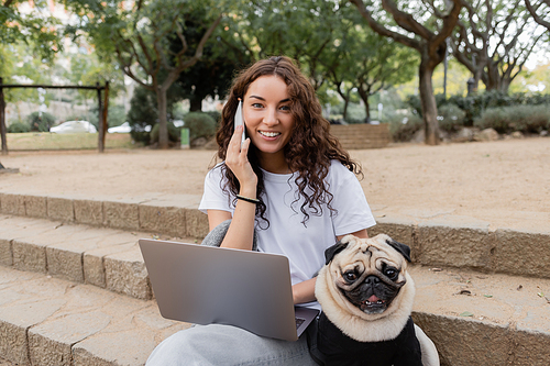 Carefree young curly woman in casual clothes looking at camera while talking on cellphone near laptop and pug dog sitting on stairs in blurred park in Barcelona, Spain