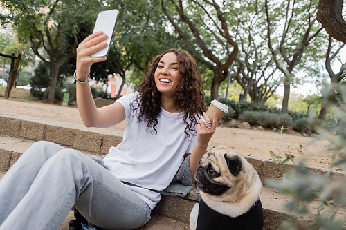 Cheerful young and curly woman in casual clothes having video call on smartphone and holding takeaway coffee near pug dog sitting on stairs in blurred park at daytime in Barcelona, Spain