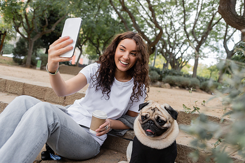 Carefree young and curly woman in casual clothes having video call on cellphone and holding coffee to go while sitting near pug dog on stairs in blurred park at daytime in Barcelona, Spain