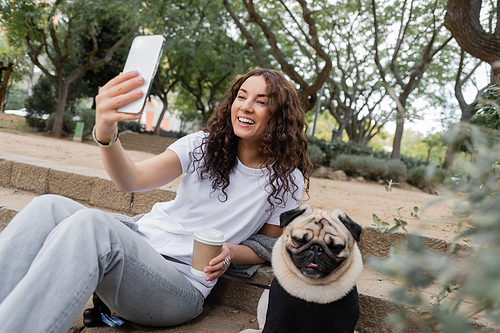 Happy young woman in casual clothes having video chat on smartphone and holding coffee to go while sitting near pug dog on stairs in blurred park at daytime in Barcelona, Spain