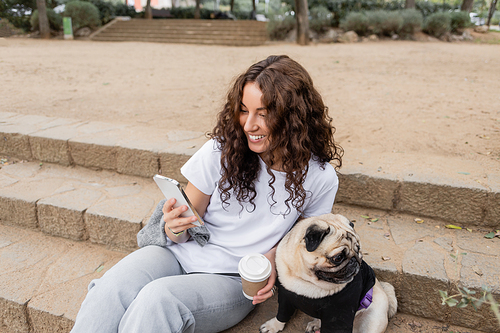 Overjoyed young curly woman in casual clothes using smartphone and holding coffee to go while sitting near pug dog on stairs in blurred park in Barcelona, Spain