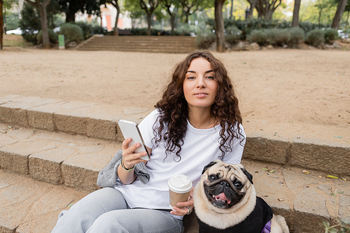 Smiling young curly woman in casual clothes using mobile phone and holding coffee to go while looking at camera near pug dog sitting on stairs in blurred park in Barcelona, Spain