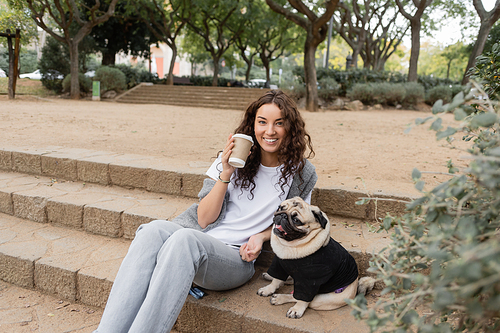Young and curly woman in casual clothes smiling at camera while holding takeaway coffee in paper cup and spending time with pug dog on stairs in park in Barcelona, Spain