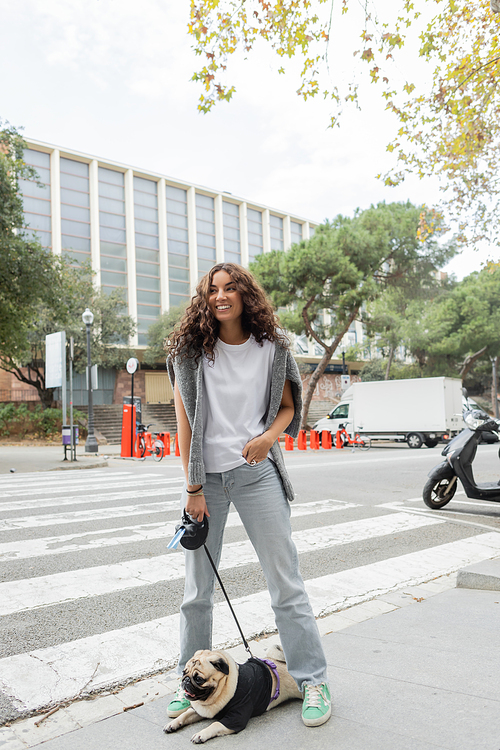 Full length of smiling young and curly woman in casual clothes holding hand in pocket of jeans and looking away while holding leash near pug dog on blurred urban street in Barcelona, Spain, building