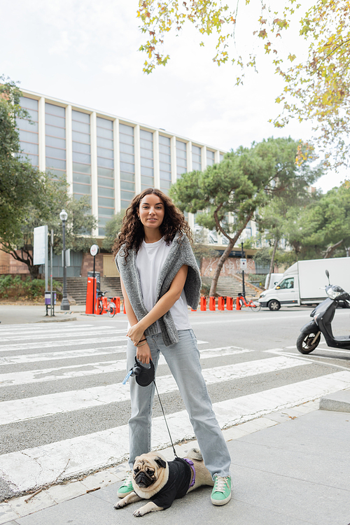 Carefree young woman in casual clothes looking at camera and holding leash while standing near pug dog near building on blurred urban street at daytime in Barcelona, Spain