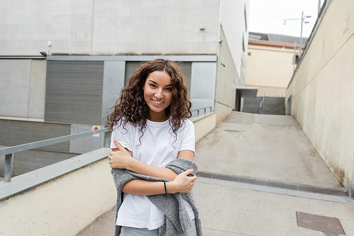 Portrait of smiling curly and pretty woman in white t-shirt and sweater looking at camera while standing near blurred industrial building at background on city street in Barcelona, Spain