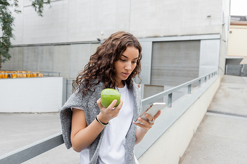 Young and pretty woman with warm sweater on shoulders using cellphone and holding fresh apple while standing on blurred urban street with industrial building in Barcelona, Spain