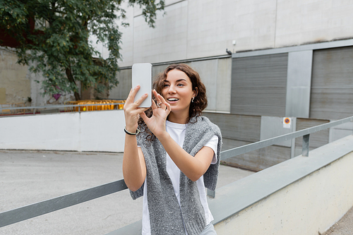 Carefree young brunette woman with warm grey sweater on shoulders having video call on smartphone while standing on urban street with industrial building at background in Barcelona, Spain