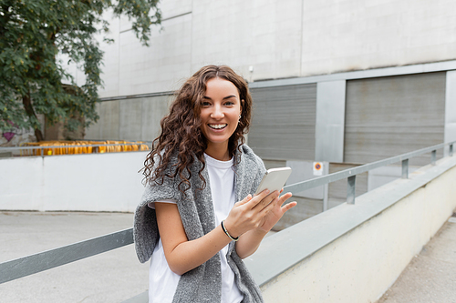 Portrait of positive and curly young woman in t-shirt and sweater looking at camera and using mobile phone while standing on city street at daytime in Barcelona, Spain, green tree