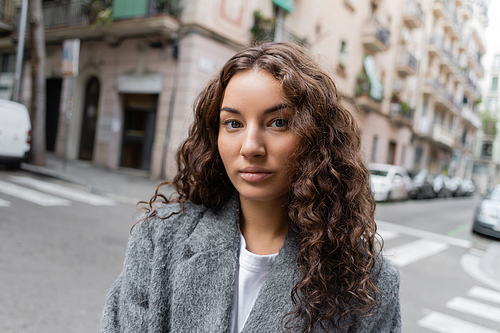 Portrait of young and curly brunette woman in casual grey jacket standing on blurred urban street with buildings at background at daytime in Barcelona, Spain