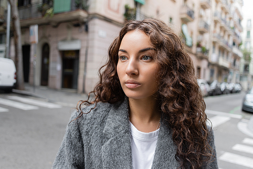 Brunette and pretty young woman in casual jacket looking away while standing on blurred city street with buildings at background in Barcelona, Spain