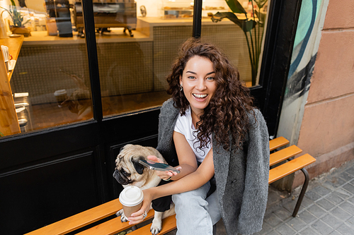 Positive young brunette woman in casual jacket using smartphone and holding coffee to go while sitting near pug dog on bench near outdoor cafe on urban street in Barcelona, Spain