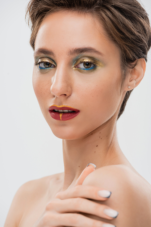 portrait of young model with bright makeup touching bare shoulder and looking at camera isolated on grey