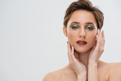 portrait of young model with bright makeup touching cheeks and looking at camera isolated on grey