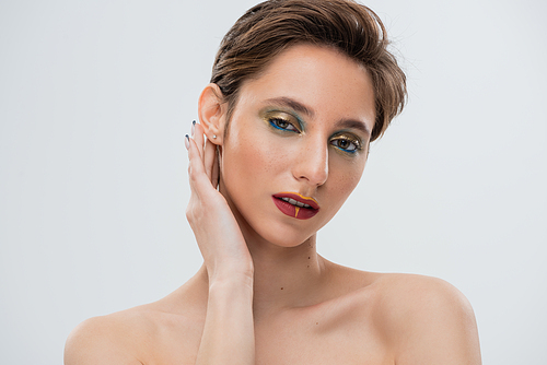 portrait of young model with bright makeup fixing shirt hair and looking at camera isolated on grey