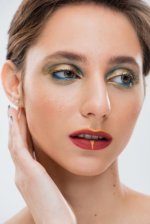 close up view of young woman with bright eye makeup fixing shirt hair and looking away isolated on grey